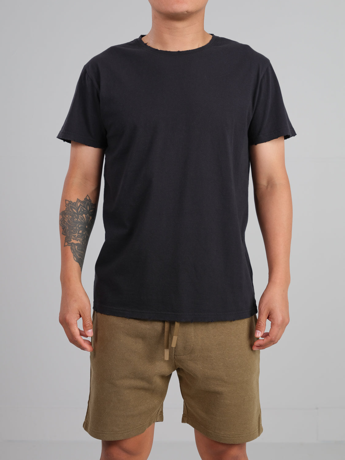 Someone in a black ID Menswear Trekker Tattered Crew Neck Tee and khaki shorts poses against a plain gray backdrop. The lightweight jersey material enhances the tattoo on their left arm. Their face is not visible.