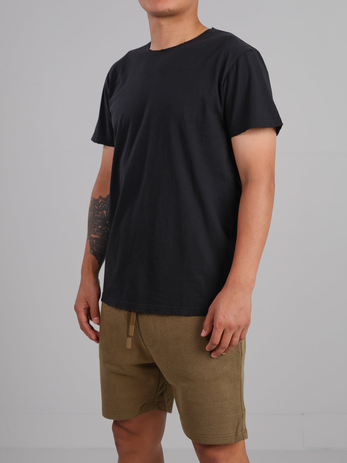 A person in an ID Menswear Trekker Tattered Crew Neck Tee and brown shorts stands against a gray background, revealing a tattoo on their left arm.
