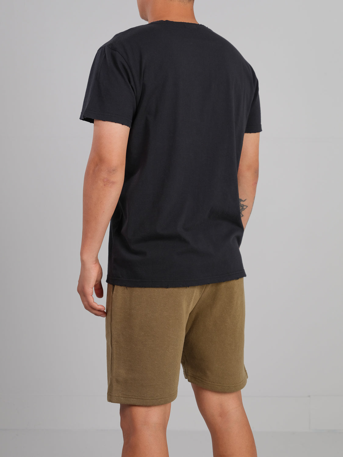 Facing away, a person dons an ID Menswear Trekker Tattered Crew Neck Tee in black and brown shorts. Part of a tattoo peeks from their arm. The lightweight jersey fabric hangs comfortably on the skin against a plain, neutral background.