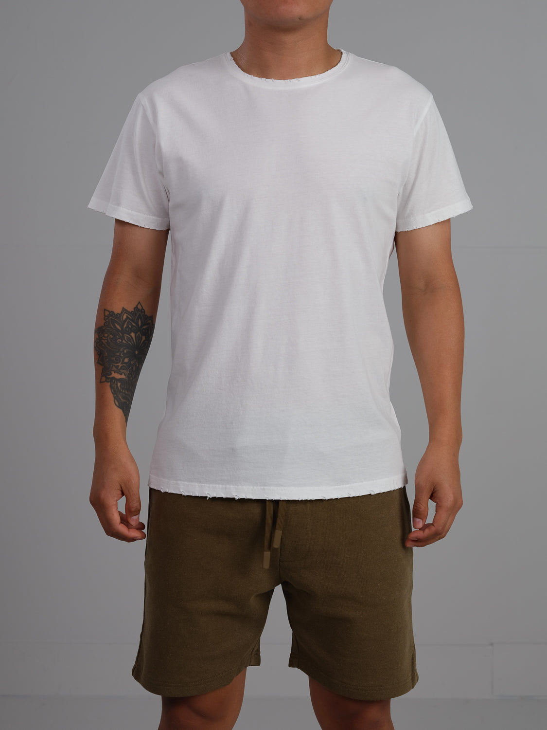 A man wearing a plain ID Menswear Trekker Tattered Crew Neck Tee and olive green shorts stands against a gray background. His lightweight jersey tee, crafted from organic cotton, complements the tattoo on his left forearm.