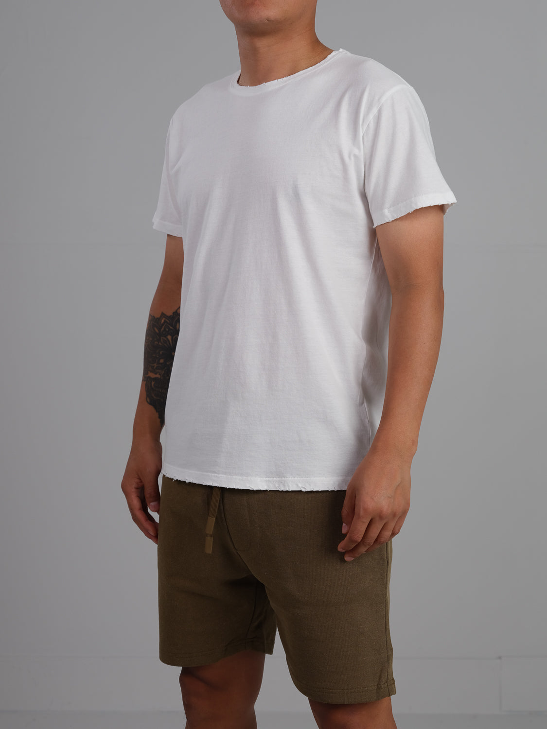 A person in an ID Menswear Trekker Tattered Crew Neck Tee and olive green shorts stands against a neutral gray backdrop. The shirts lightweight jersey accents their short hair and forearm tattoo, with their body slightly angled to the left.