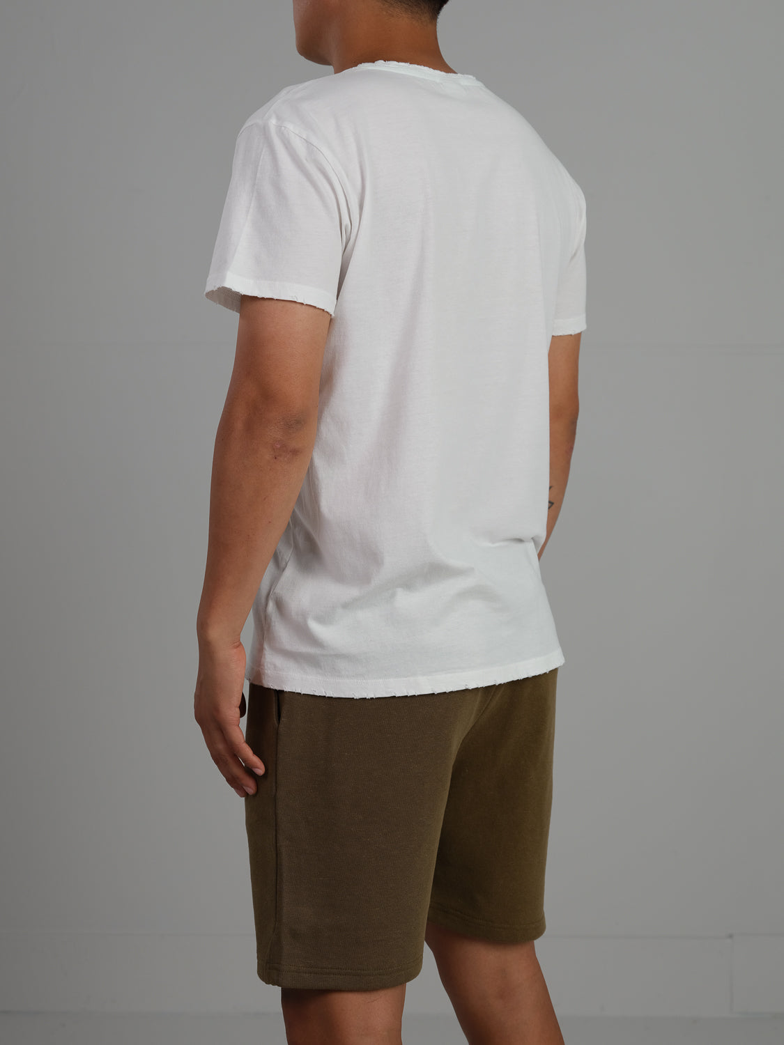 A person stands facing away in an ID Menswear Trekker Tattered Crew Neck Tee and olive green shorts. The lightweight jersey fabric drapes casually against the plain gray backdrop, with their arms hanging relaxed by their sides.