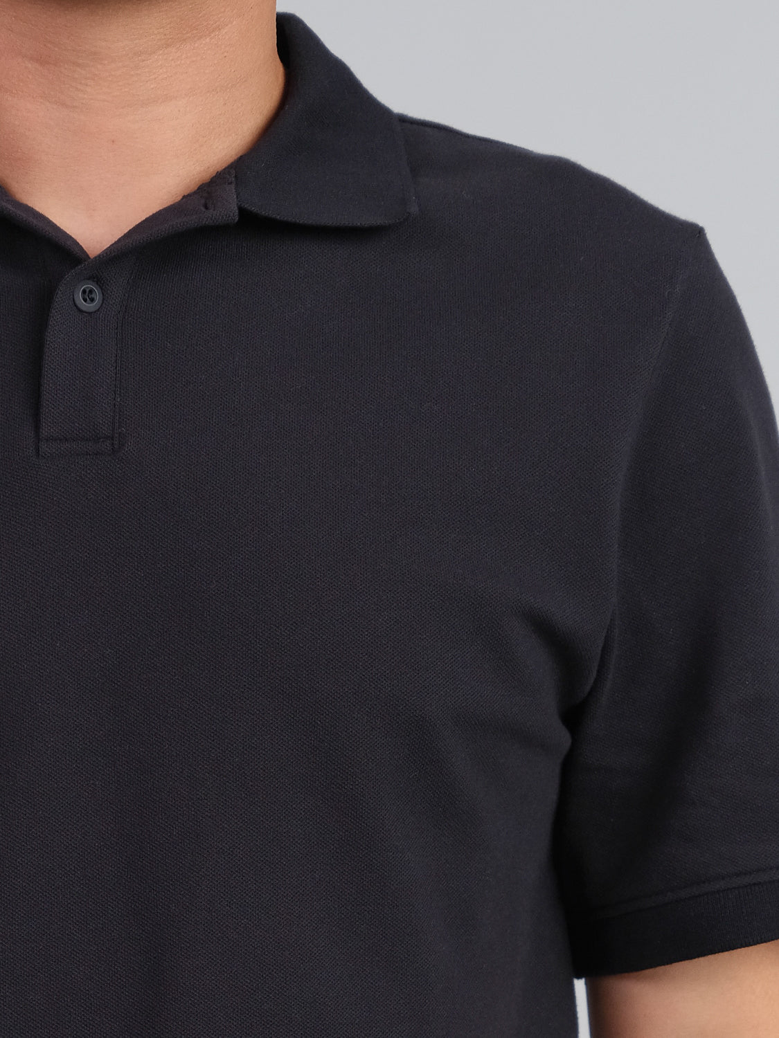 Close-up of someone wearing a black ID Menswear Ranger Classic Cotton Mesh Polo. The focus is on the upper half, featuring a classic collar and one visible button, crafted from lightweight cotton. The background is plain light gray.