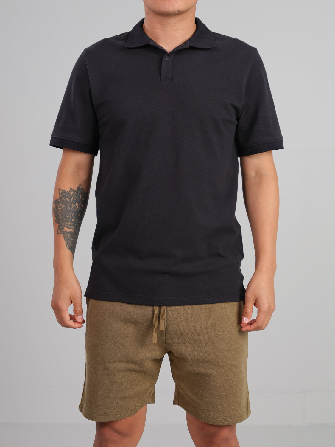 A person in a black ID Menswear Ranger Classic Cotton Mesh Polo and khaki shorts stands against a plain background. The lightweight polo complements the classic style, while their left arm shows a tattooed geometric pattern.
