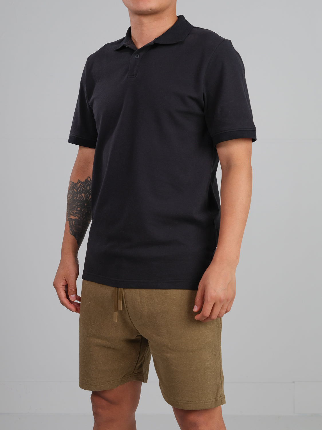 A person wearing an ID Menswear Ranger Classic Cotton Mesh Polo in black and khaki shorts stands against a plain gray background. Crafted from lightweight cotton, their attire complements the tattoo on their left forearm. The image does not show their head.