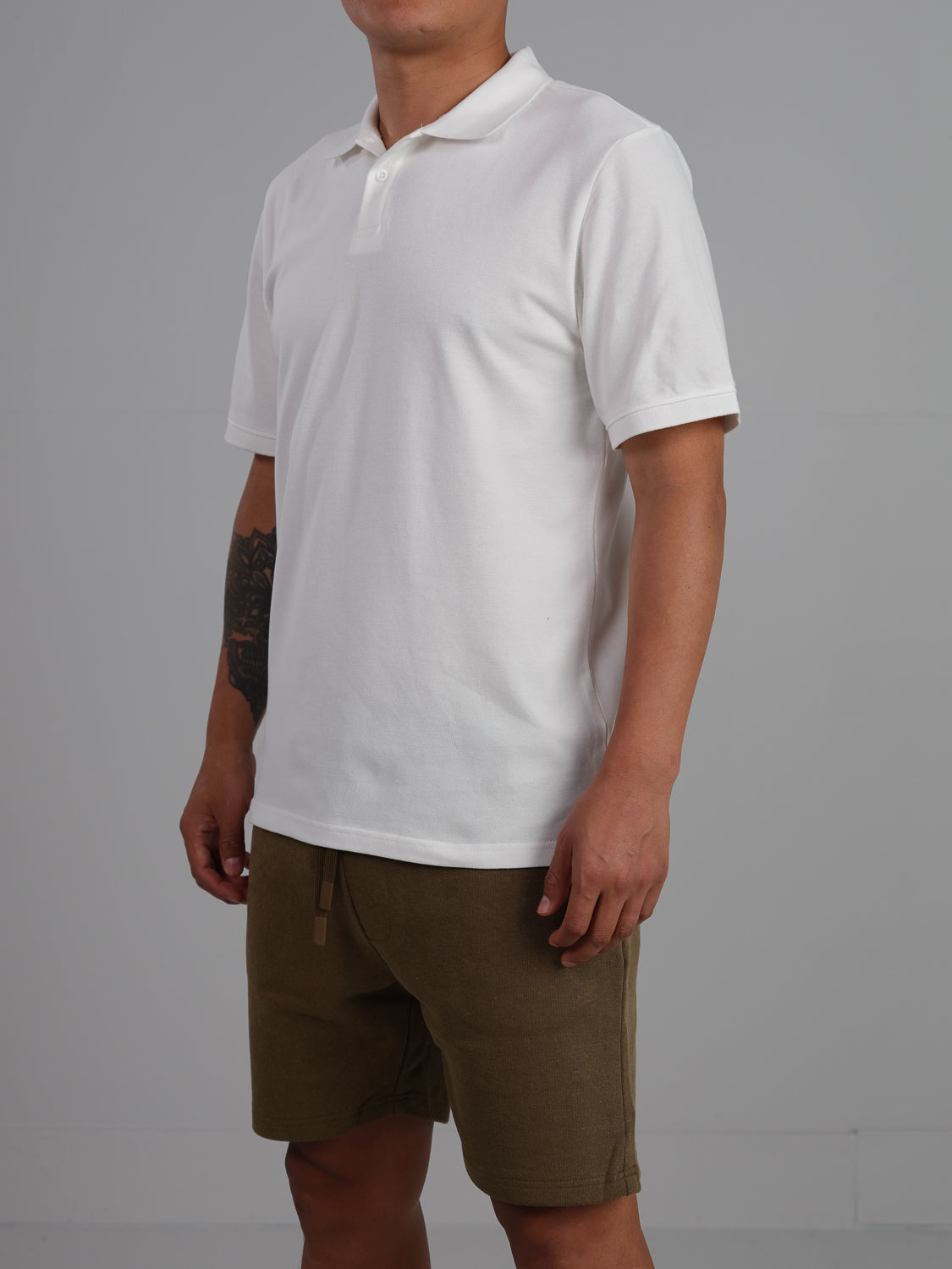 A person models an ID Menswear Ranger Classic Cotton Mesh Polo and olive green shorts against a gray backdrop. The lightweight cotton shirt enhances the relaxed look. A tattoo decorates their left arm, while their face is hidden from view.