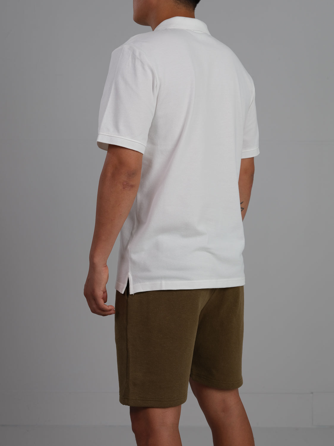 In front of a gray backdrop, an individual wears ID Menswears Ranger Classic Cotton Mesh Polo in white paired with olive green shorts. The lightweight ensemble offers comfort and elegance as the person faces away from the camera, keeping their identity hidden.