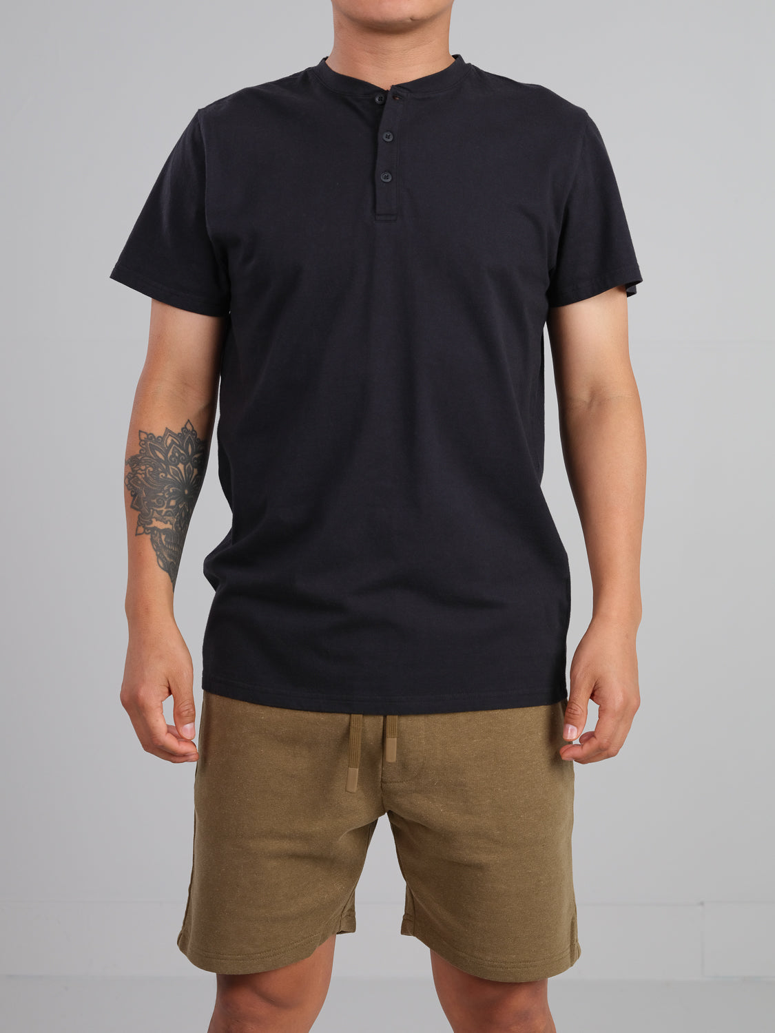 A person in a black Champion Organic Cotton Henley by ID Menswear with tan shorts, sporting a forearm tattoo, stands against a plain gray background, showcasing a classic look.