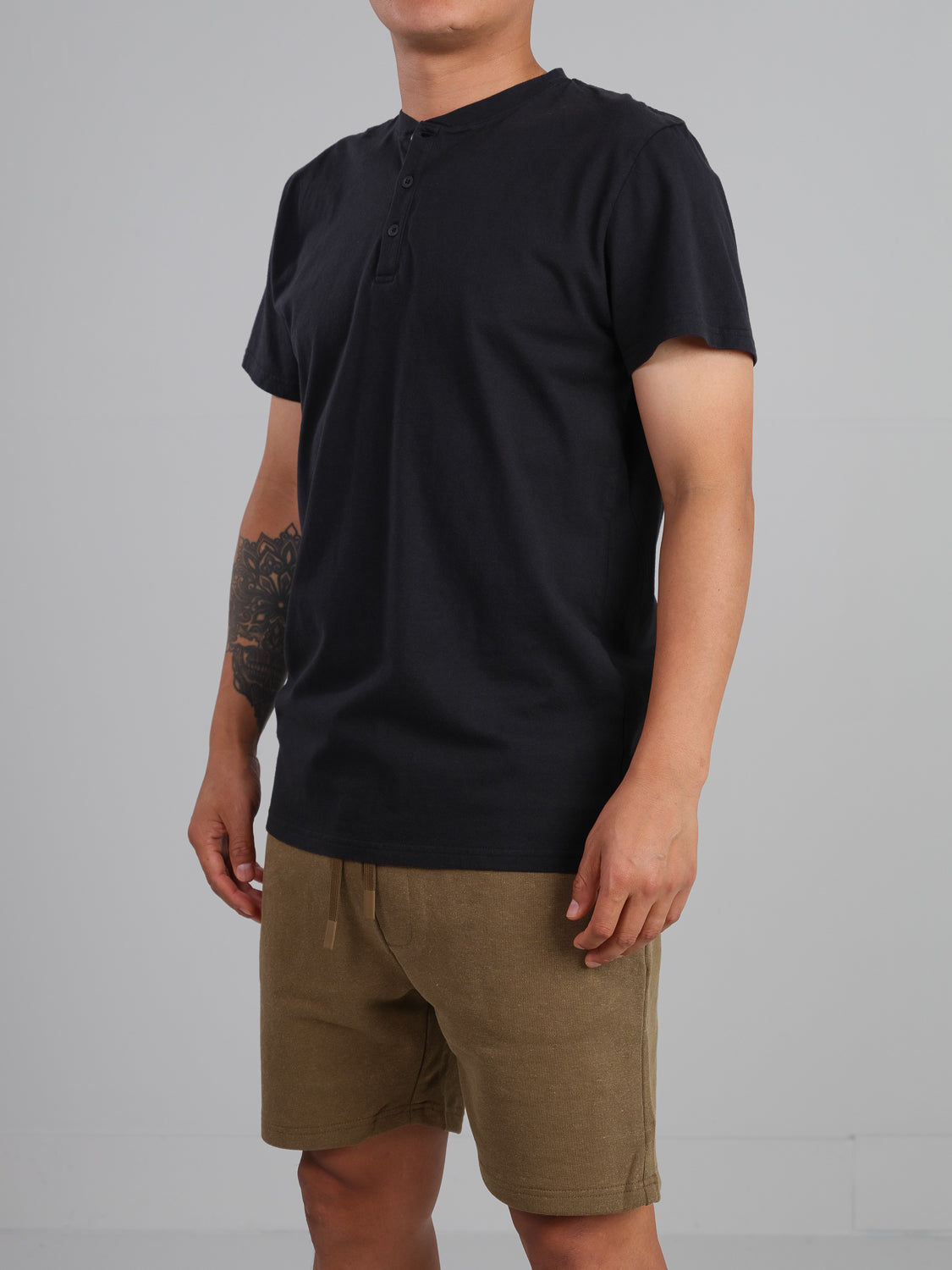 A person models the Champion Organic Cotton Henley by ID Menswear, showcasing a classic silhouette in black with a short-sleeve design, paired with tan shorts. They stand against a gray background, tattooed on their left arm while facing slightly to the side.