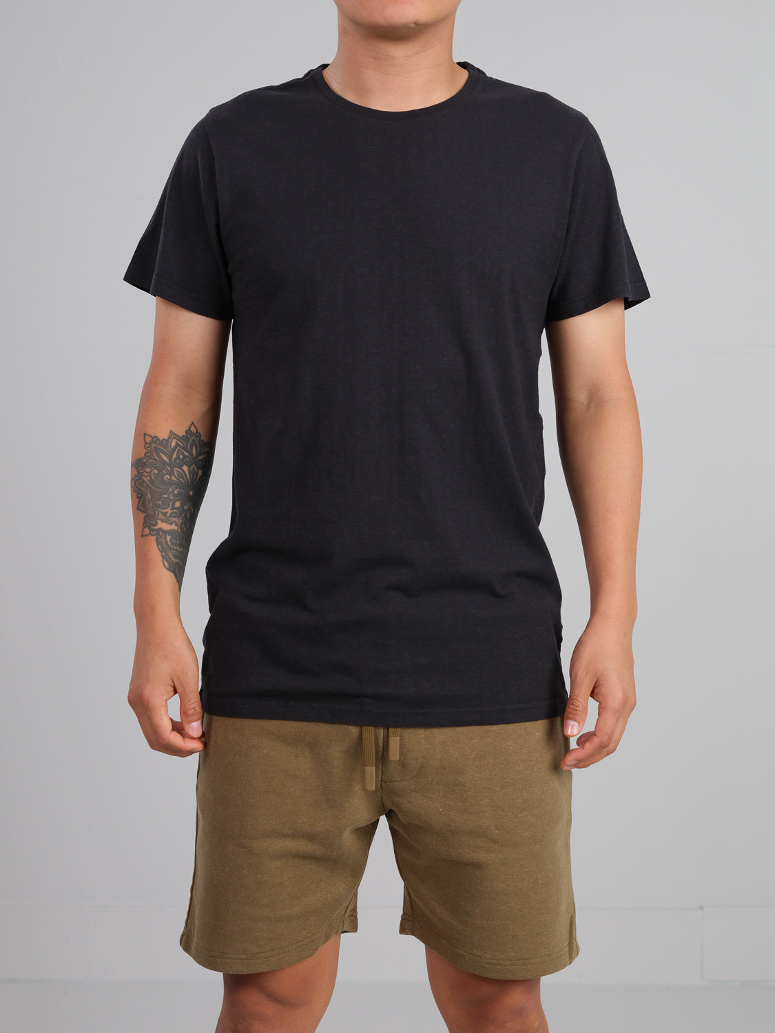 A person wearing the Dope Hemp Organic Cotton Tee by ID Menswear and beige shorts, both crafted from sustainable fabric, stands against a gray background. Their arms are relaxed at their sides, one displaying a tattoo. The image highlights their torso and eco-friendly casual attire.