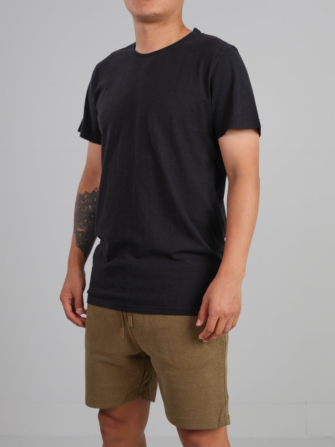 Against a gray backdrop, a person wears the Dope Hemp Organic Cotton Tee by ID Menswear and khaki shorts. The focus is on their tattooed left forearm and sustainable fabric choice.