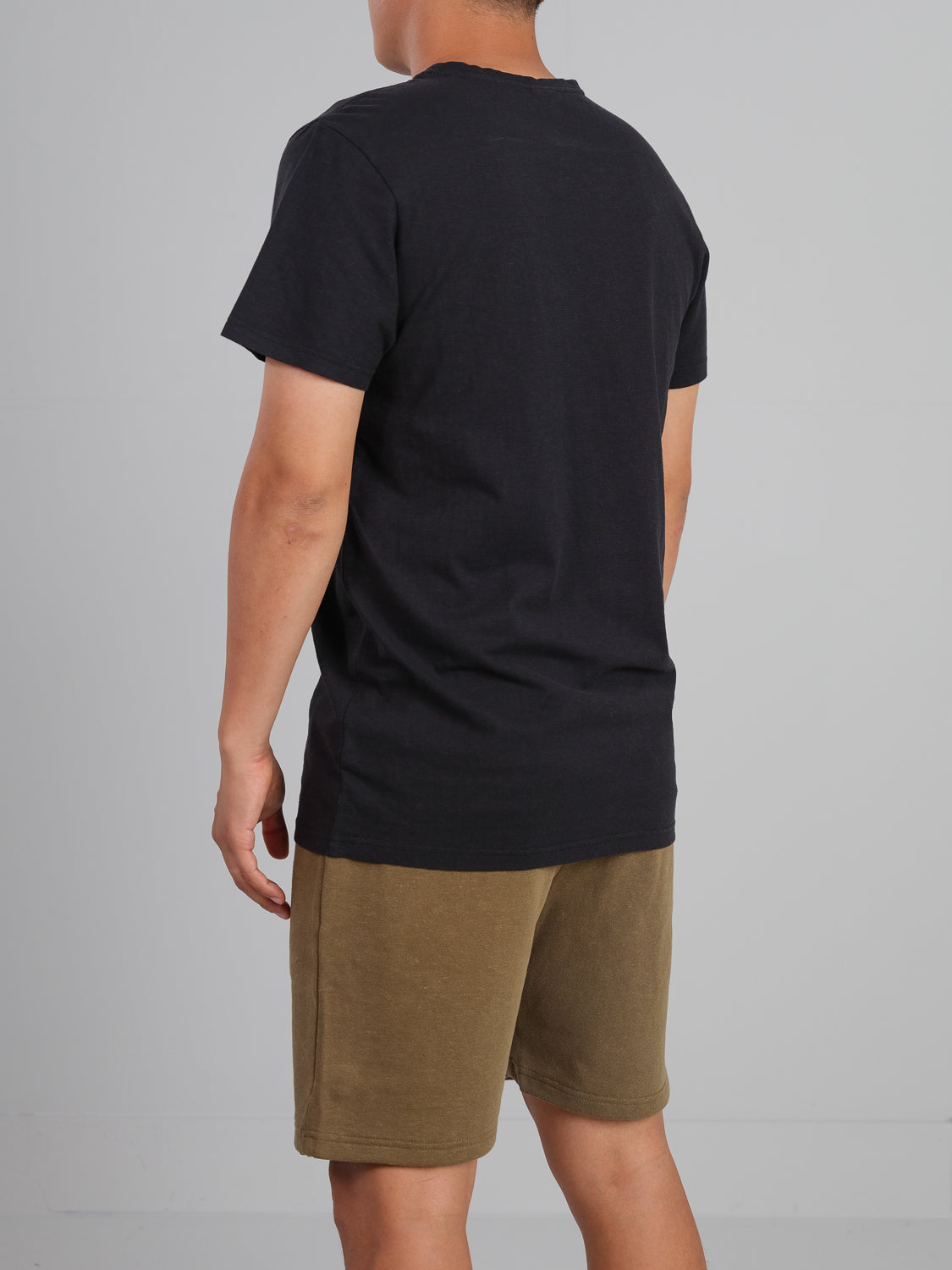 A person in a Dope Hemp Organic Cotton Tee by ID Menswear and olive green shorts stands against a gray wall, highlighting their sustainable fabric choices.