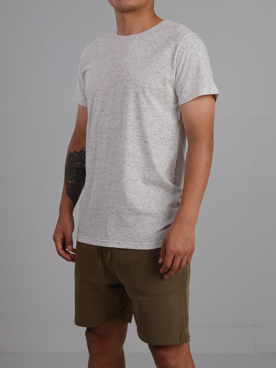 A person wears the Outclass Speckled Crew Neck Tee from ID Menswear in light gray and olive green shorts, standing against a plain background. Their tattooed right arm is visible while their face is out of frame, and the cotton/poly jersey fabric ensures comfort and style.