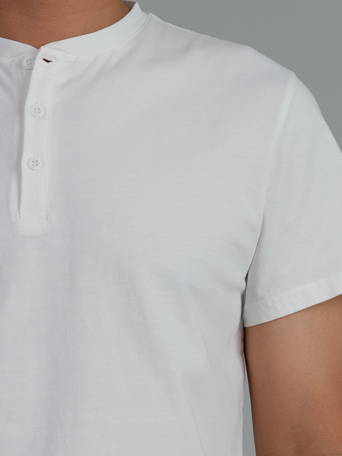 A close-up shows an individual wearing the Champion Organic Cotton Henley by ID Menswear, a white jersey with short sleeves, round neckline, and three-button placket against a plain gray background. Only the upper body is visible.