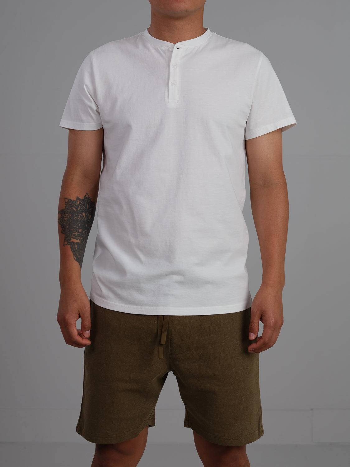 Against a plain gray background, an individual is seen wearing ID Menswears Champion Organic Cotton Henley, featuring short sleeves, paired with olive green drawstring shorts. A tattoo on their right forearm enhances the classic silhouette of this attire crafted from organic cotton jersey.