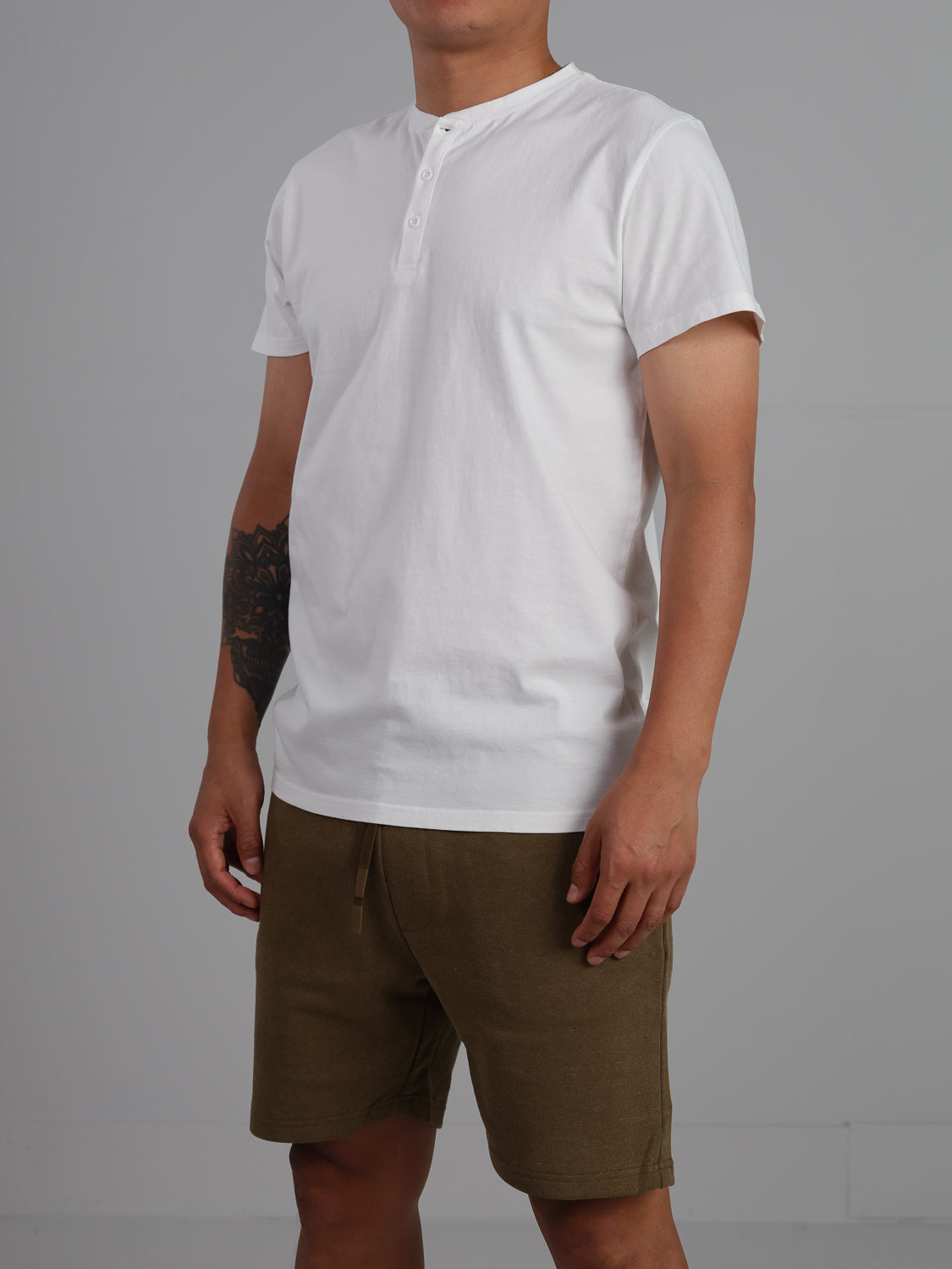 Against a neutral background, someone wears ID Menswears Champion Organic Cotton Henley—a white short-sleeve shirt made from organic cotton jersey—paired with olive green shorts, perfectly highlighting their arm tattoo.