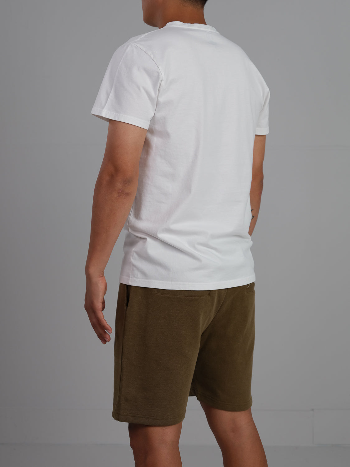 A person in a classic silhouette, wearing an ID Menswear Champion Organic Cotton Henley and olive green shorts, stands against a gray background. Their arms rest casually by their sides as the image captures them from the back.