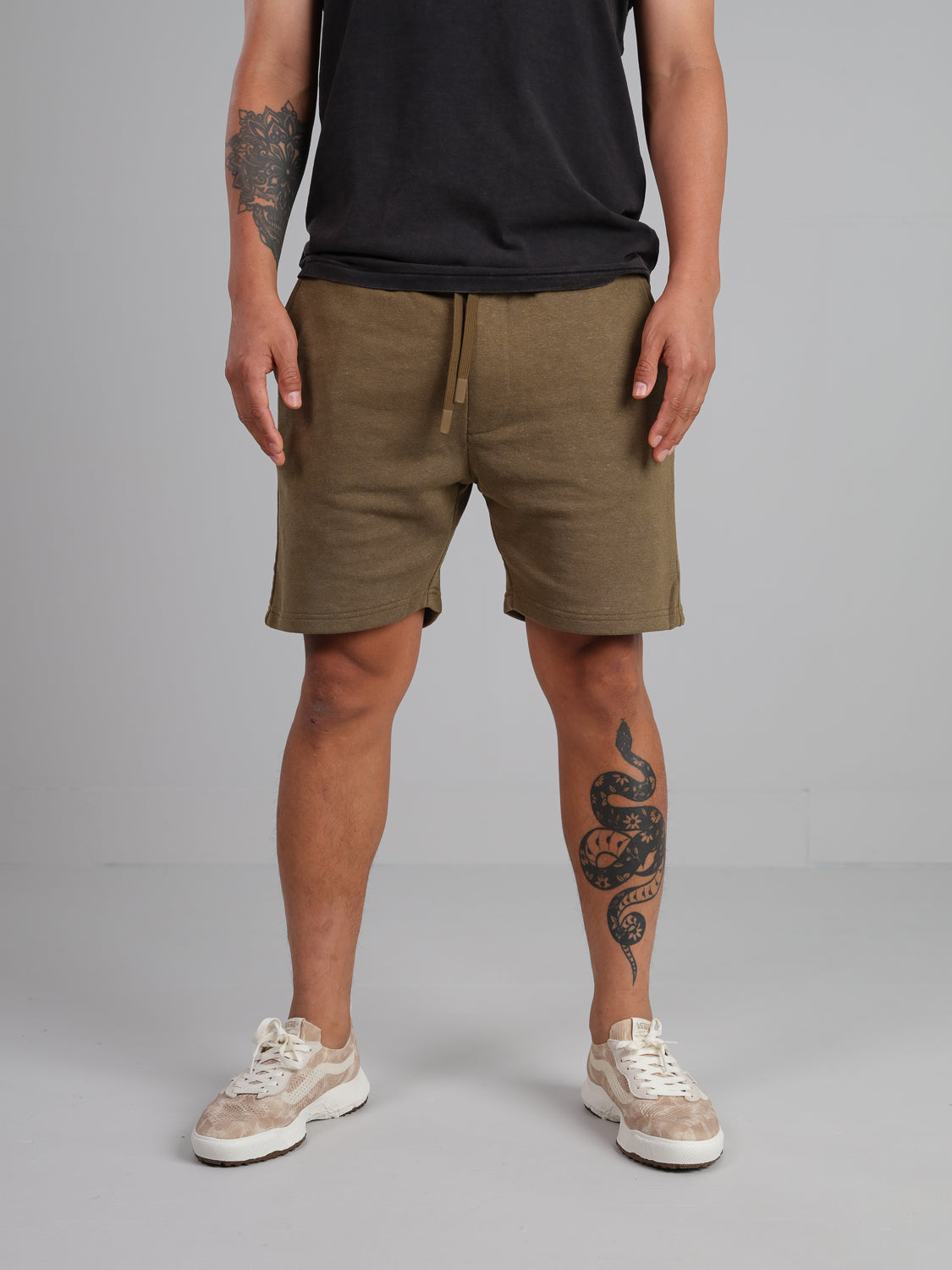 A man wears ID Menswears Walker Hemp Cotton Terry Shorts and a black t-shirt, standing neutrally. His legs and left forearm feature tattoos. Hes wearing white and beige sneakers against a plain light gray backdrop.