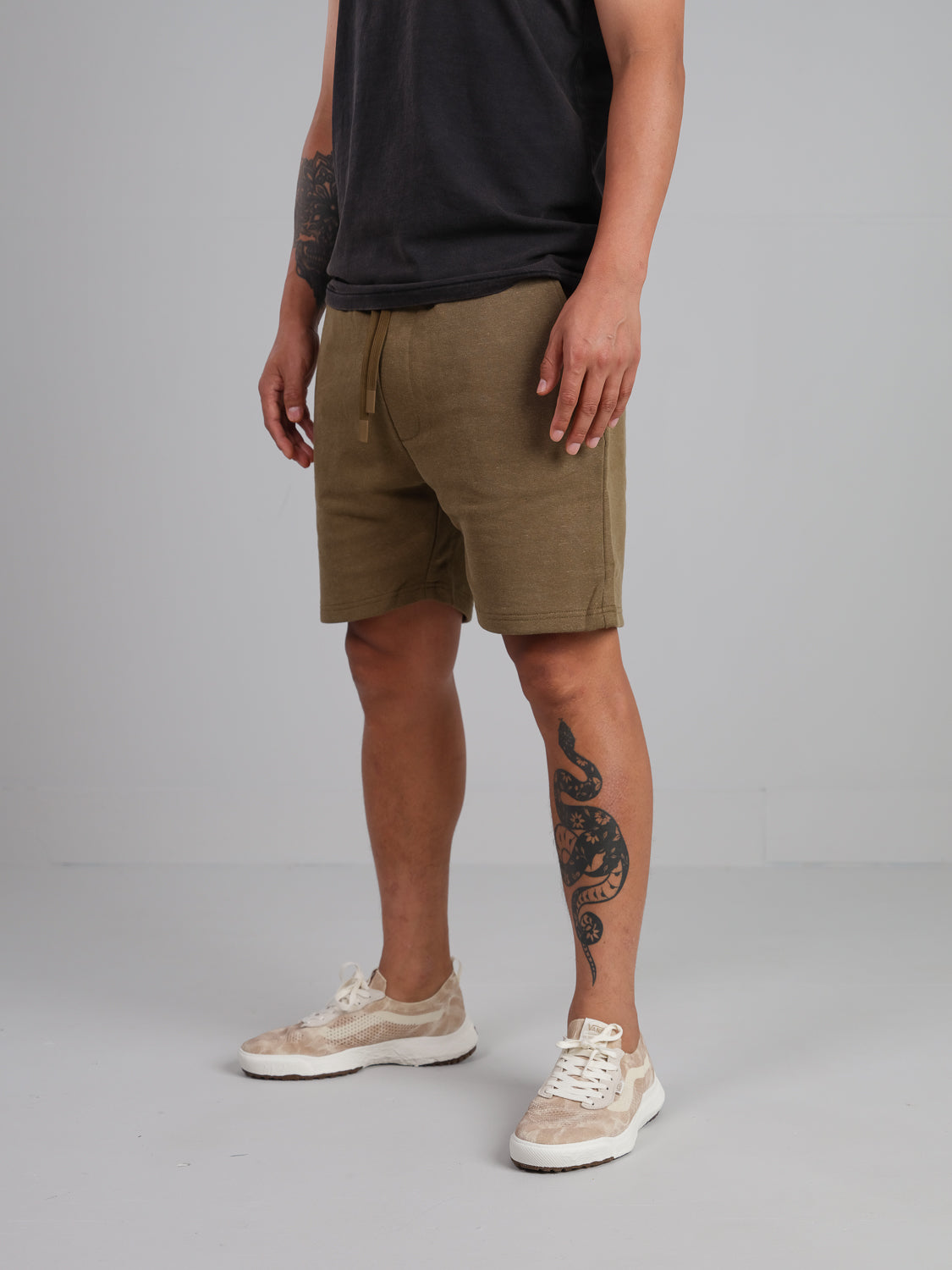 A person wearing a black shirt and ID Menswears Walker Hemp Cotton Terry Shorts in olive stands in a minimalistic room. They sport beige sneakers and have a snake tattoo on their left leg. The floor and walls are gray.
