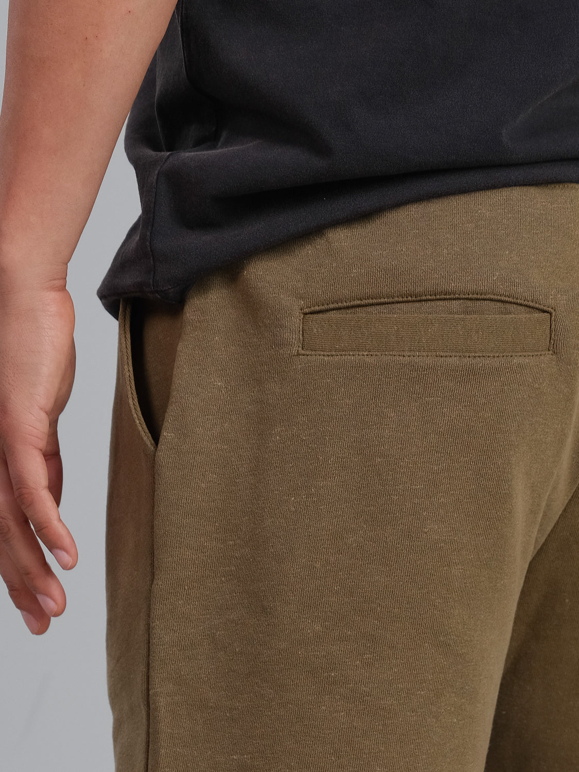 Close-up of a person wearing ID Menswears Walker Hemp Cotton Terry Short in brown. The focus is on the side and back pocket area, showcasing visible seams with a relaxed hand resting beside them against a plain gray background.