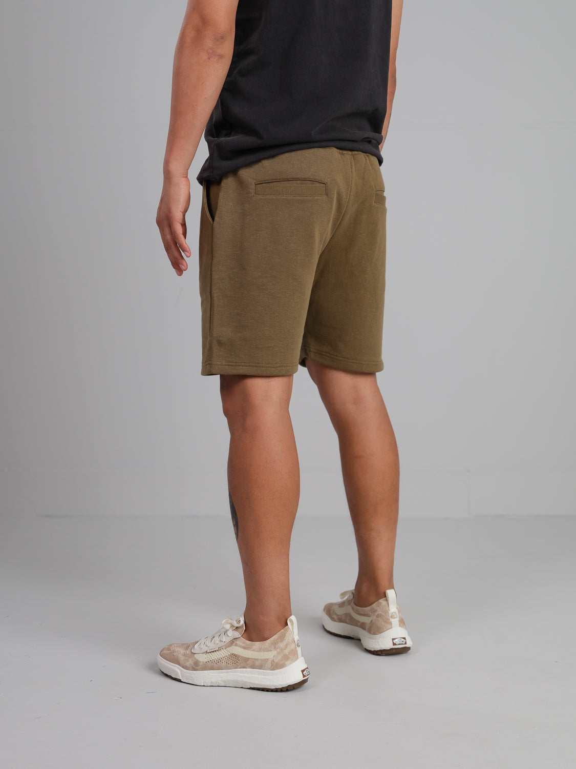 Against a plain gray background, someone in a black shirt faces away, highlighting the back of casual olive green Walker Hemp Cotton Terry Shorts from ID Menswear and wearing beige and white sneakers.