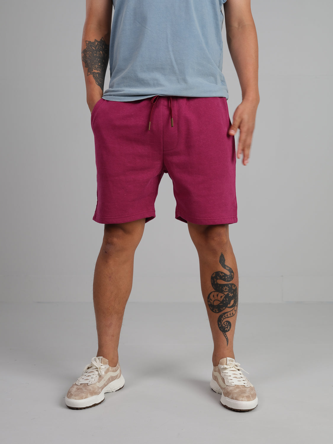 A person in a light blue t-shirt, Walker Hemp Cotton Terry Shorts from ID Menswear, and beige sneakers stands against a plain background. Their left leg displays a tattoo of a coiled snake.