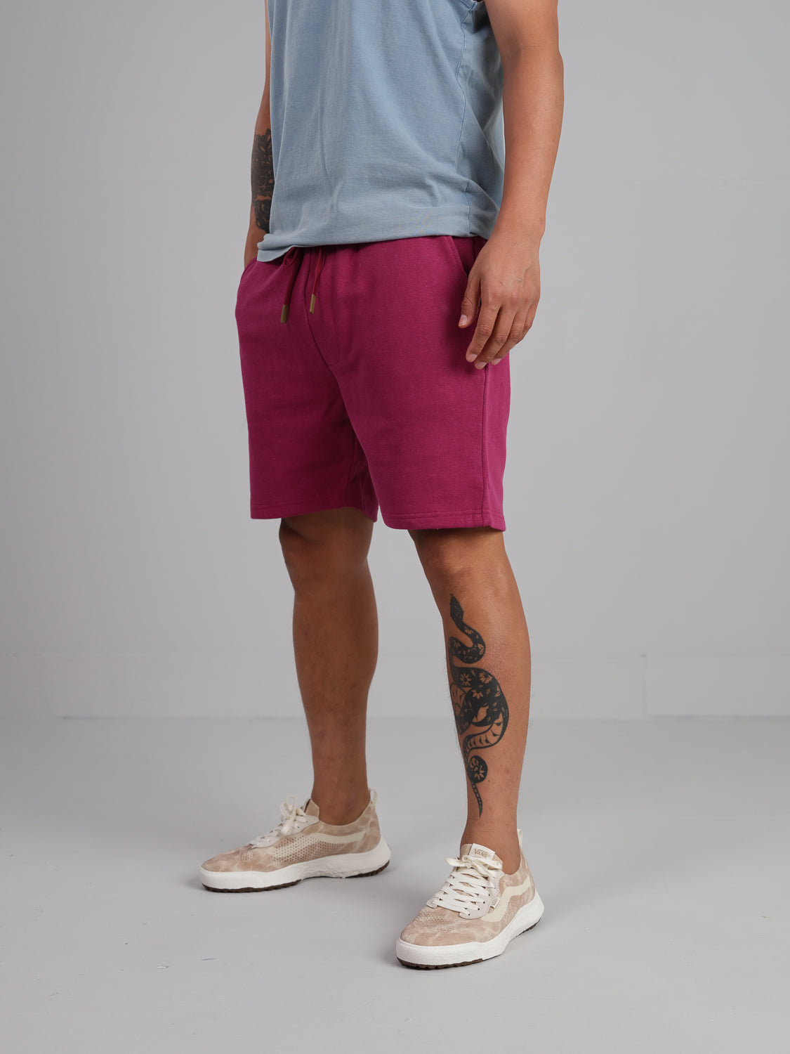 A person wearing a light blue t-shirt and Walker Hemp Cotton Terry Short by ID Menswear stands with their right hand in their pocket. They have a tattoo on their right leg and are wearing white sneakers with beige accents, set against a plain, light gray wall.