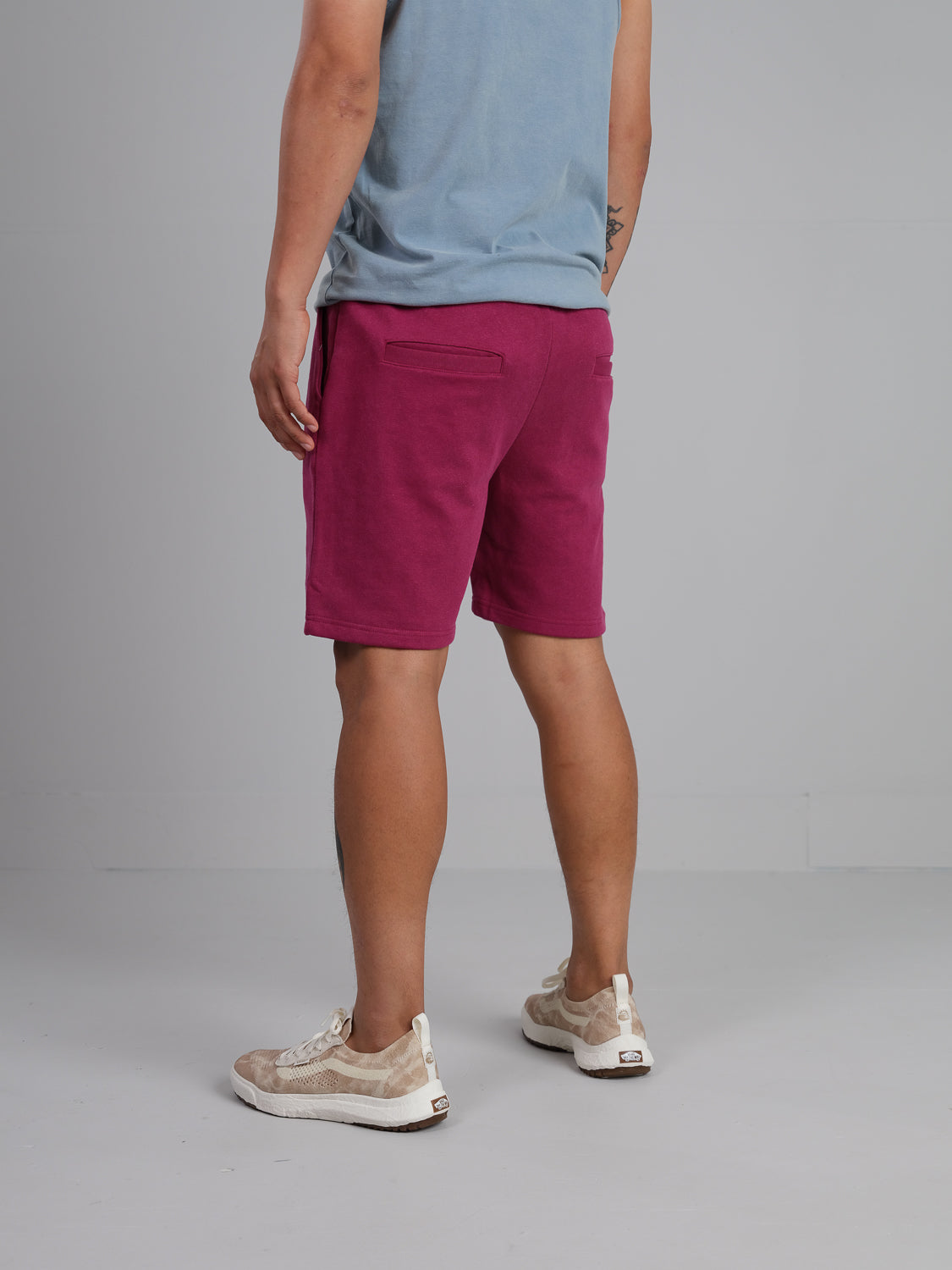 A person faces away, wearing an ID Menswear blue shirt with Walker Hemp Cotton Terry Shorts in a pinkish-red color and beige sneakers. Against a plain light gray wall and floor, their right arm showcases an intricate tattoo.