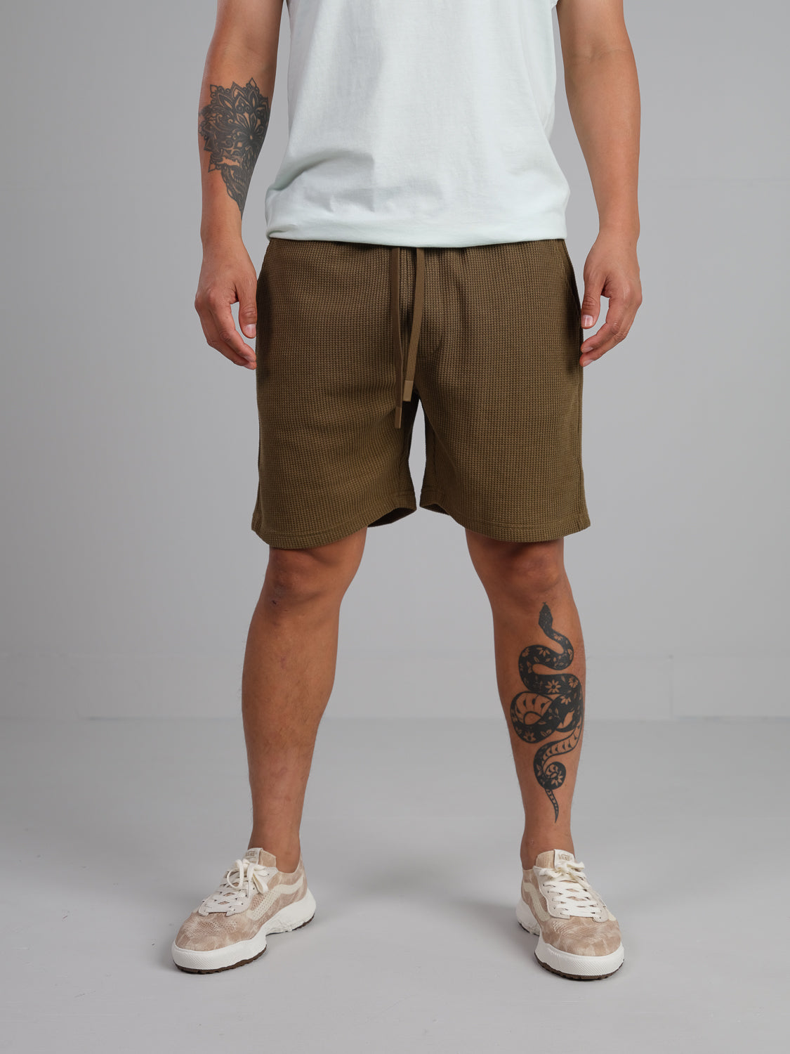 A person dons a white tee, Walker Waffle Knit Short by ID Menswear, and white sneakers against a neutral backdrop. A floral tattoo adorns their left arm, and a snake design embellishes their right leg.