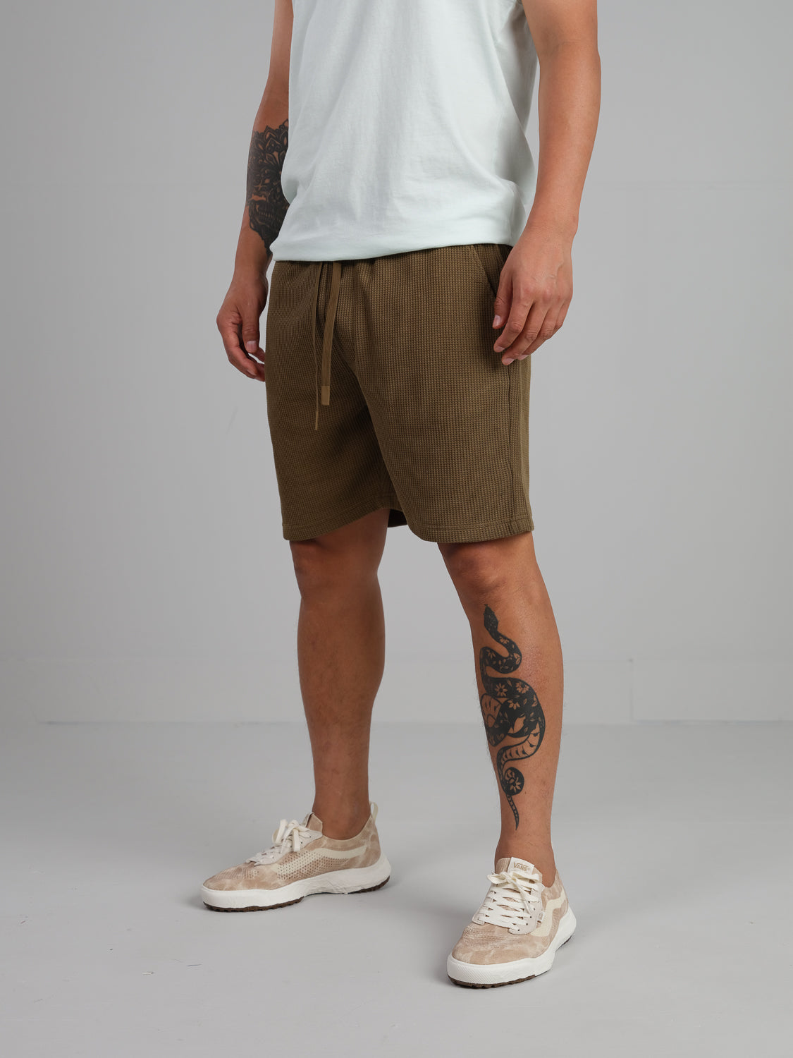 A person stands wearing a white t-shirt, Walker Waffle Knit Short by ID Menswear, and beige sneakers. They have tattoos on their left arm and right calf against a plain, light-colored wall and floor.