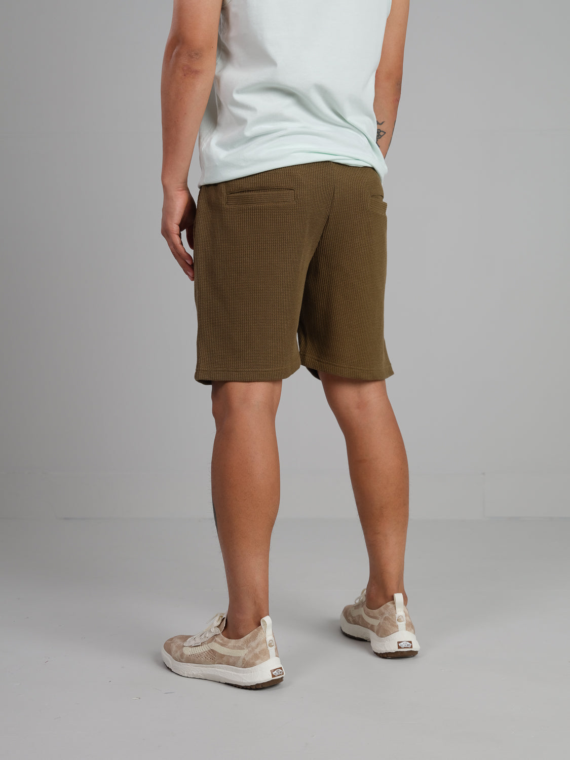 A person stands in a neutral space wearing a light shirt, Walker Waffle Knit Short from ID Menswear in olive green, and beige sneakers against a plain gray background.