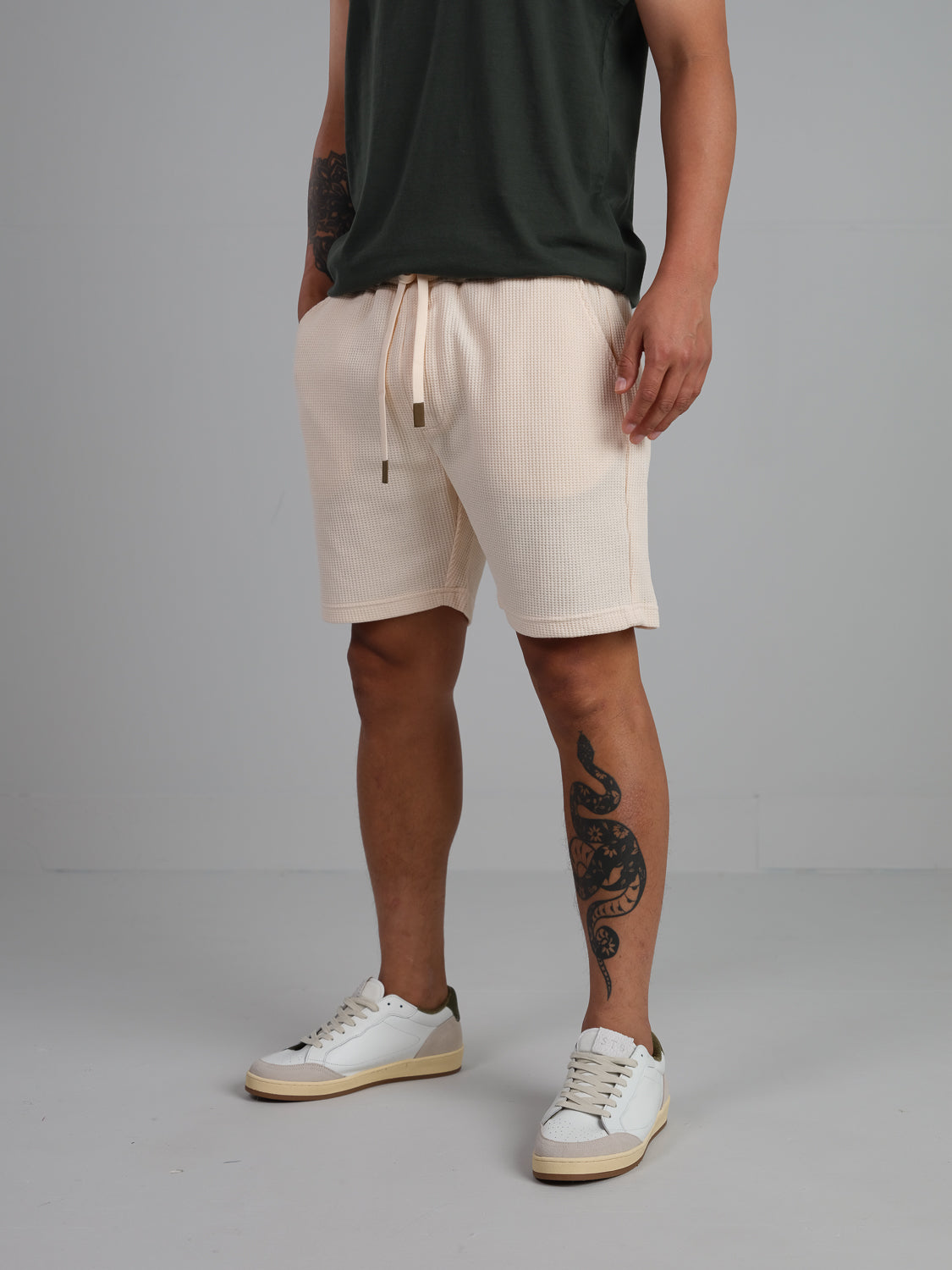 An individual sports a dark green t-shirt, ID Menswears Walker Waffle Knit Shorts in beige, and white sneakers with green details. A snake tattoo adorns their leg against a plain gray backdrop.