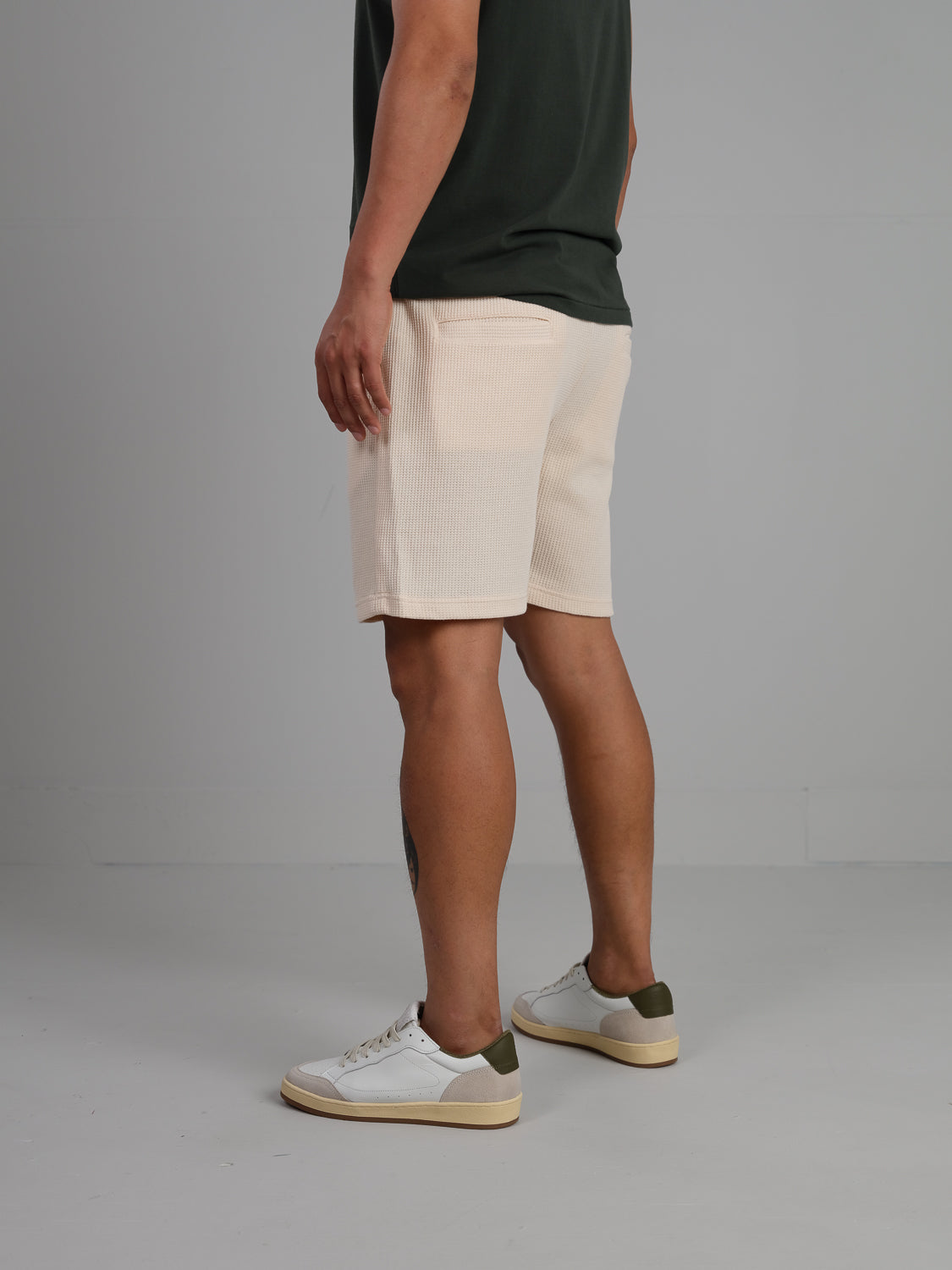 A person stands sideways, elegantly wearing an ID Menswear dark green T-shirt, Walker Waffle Knit Shorts in beige, and white sneakers with green accents, all against a plain light gray background.