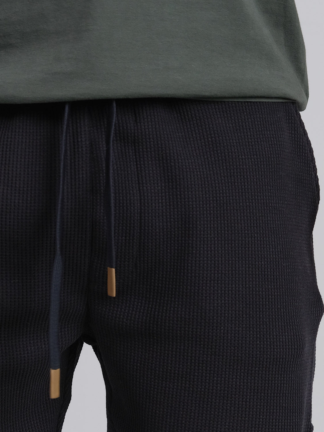 A person is wearing a textured dark pair of drawstring Walker Waffle Knit Shorts by ID Menswear, paired with a dark green shirt. The focus is on the midsection, highlighting the shorts waistband and drawstring.