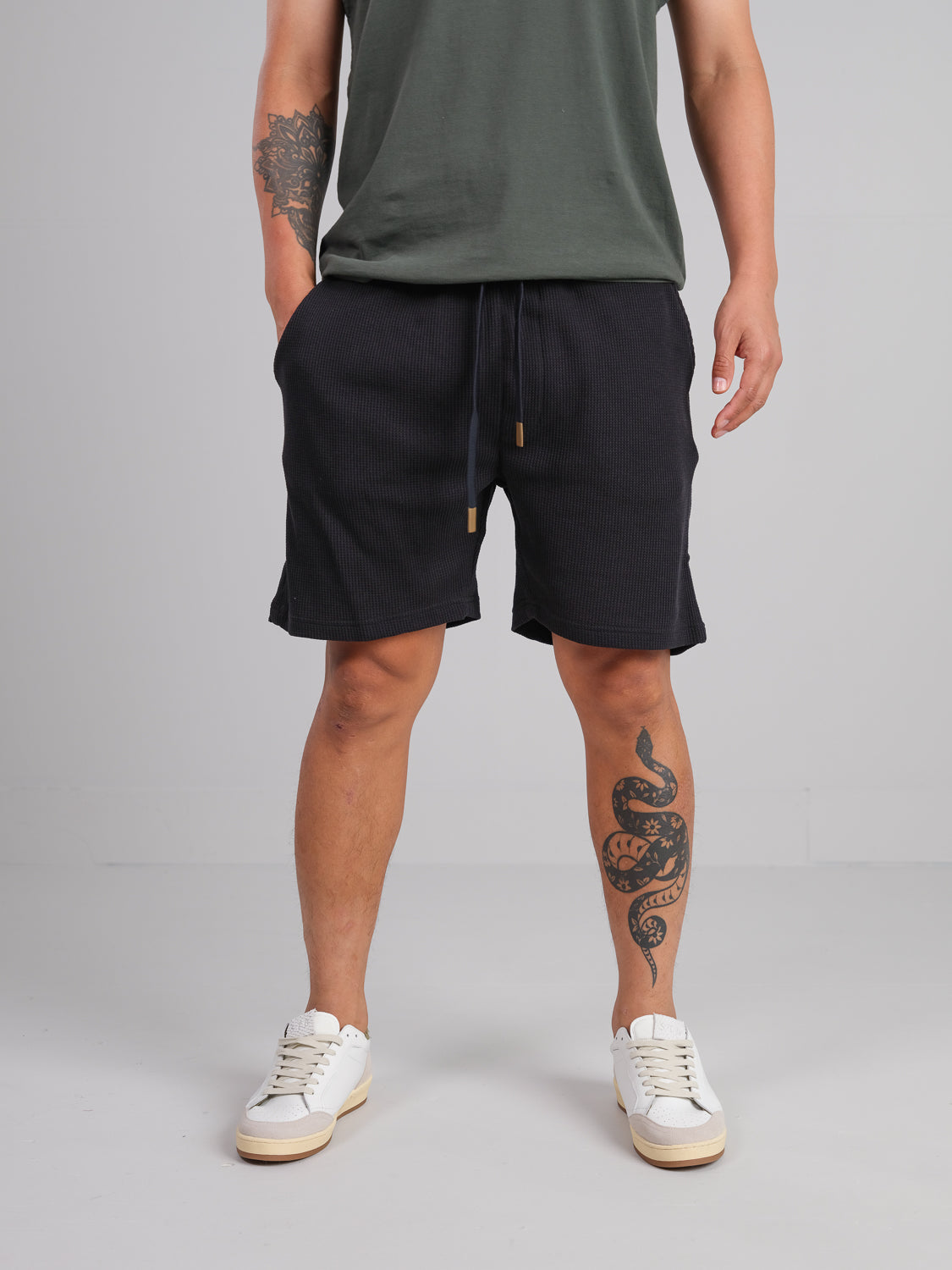 A person wearing a green T-shirt, ID Menswear Walker Waffle Knit Short in black, and white sneakers is standing. They have a coiled snake tattoo on their left leg and another tattoo on their left arm against a plain gray background.