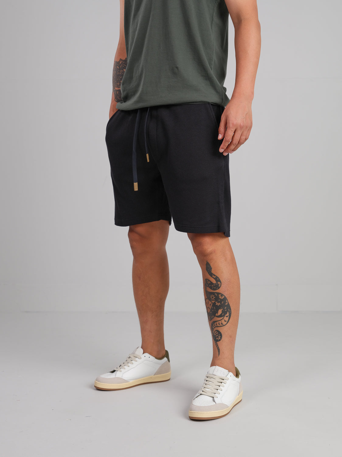 A person stands wearing a dark green t-shirt, Walker Waffle Knit Short in black by ID Menswear, and white sneakers with green accents. They have a tattoo on their left calf against a plain light gray background.