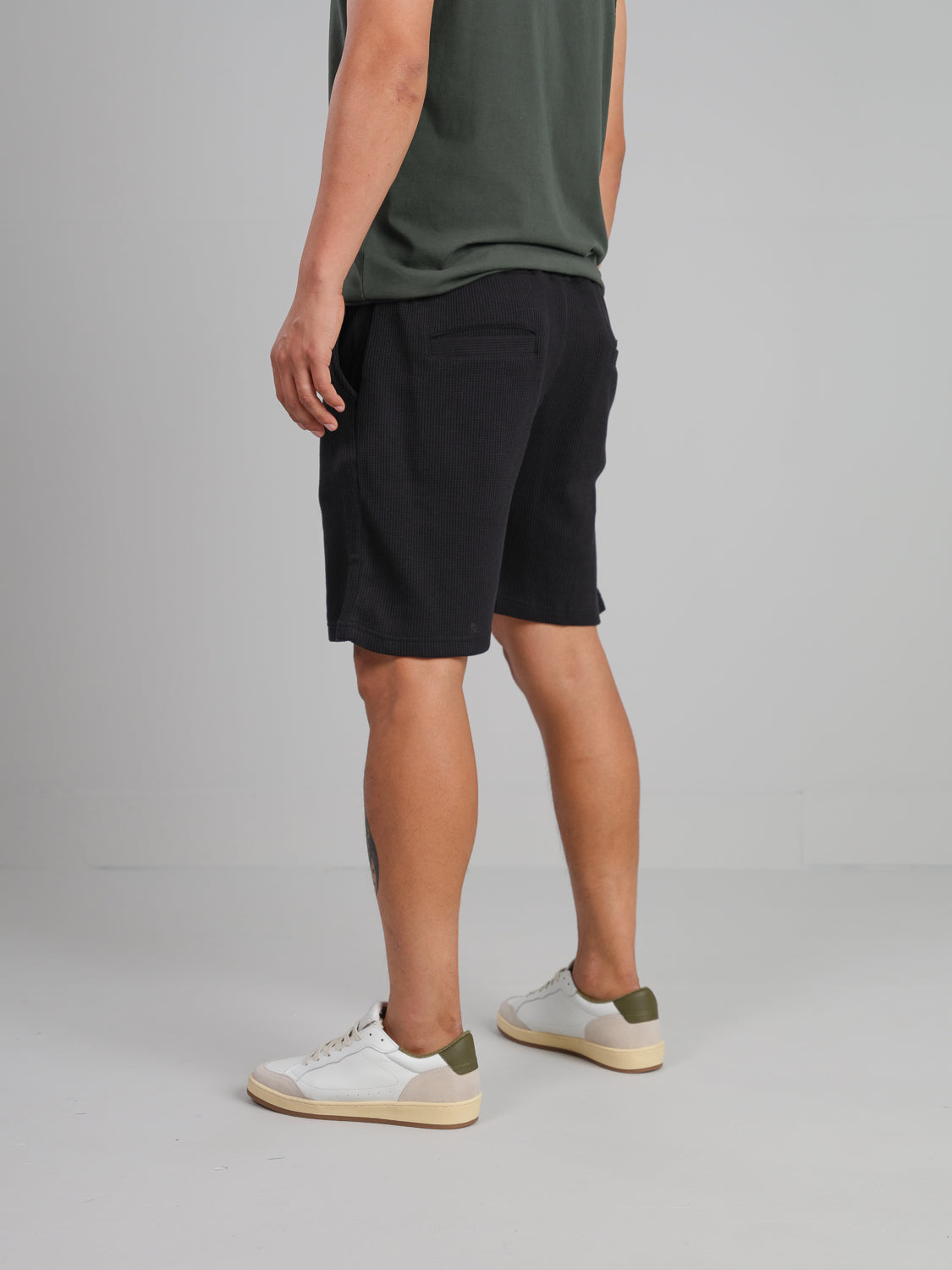 A person stands facing a light gray wall, wearing a green shirt, Walker Waffle Knit Short in black by ID Menswear, and white sneakers with green accents. The focus is on their lower body and legs.