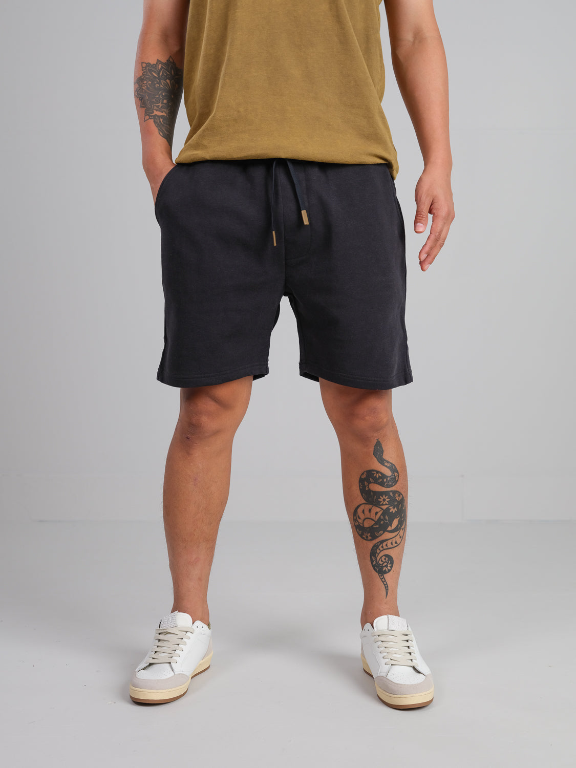 A person in a mustard organic cotton shirt, ID Menswears Walker Hemp Cotton Terry Short, and white sneakers stands with a hand in their pocket. A snake tattoo is visible on their left leg against a plain gray background.
