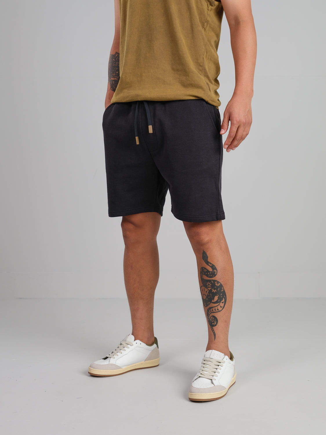 A person in a mustard organic cotton t-shirt and ID Menswears black Walker Hemp Cotton Terry Short, paired with white sneakers, stands against a plain background. One leg displays a dragon tattoo, while their face is not visible.