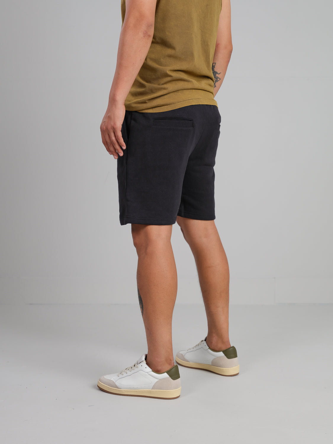 A person in a brown shirt and Walker Hemp Cotton Terry Shorts by ID Menswear stands sideways, tattoos on their right arm and left calf. They wear white sneakers with green accents against a plain light gray background.