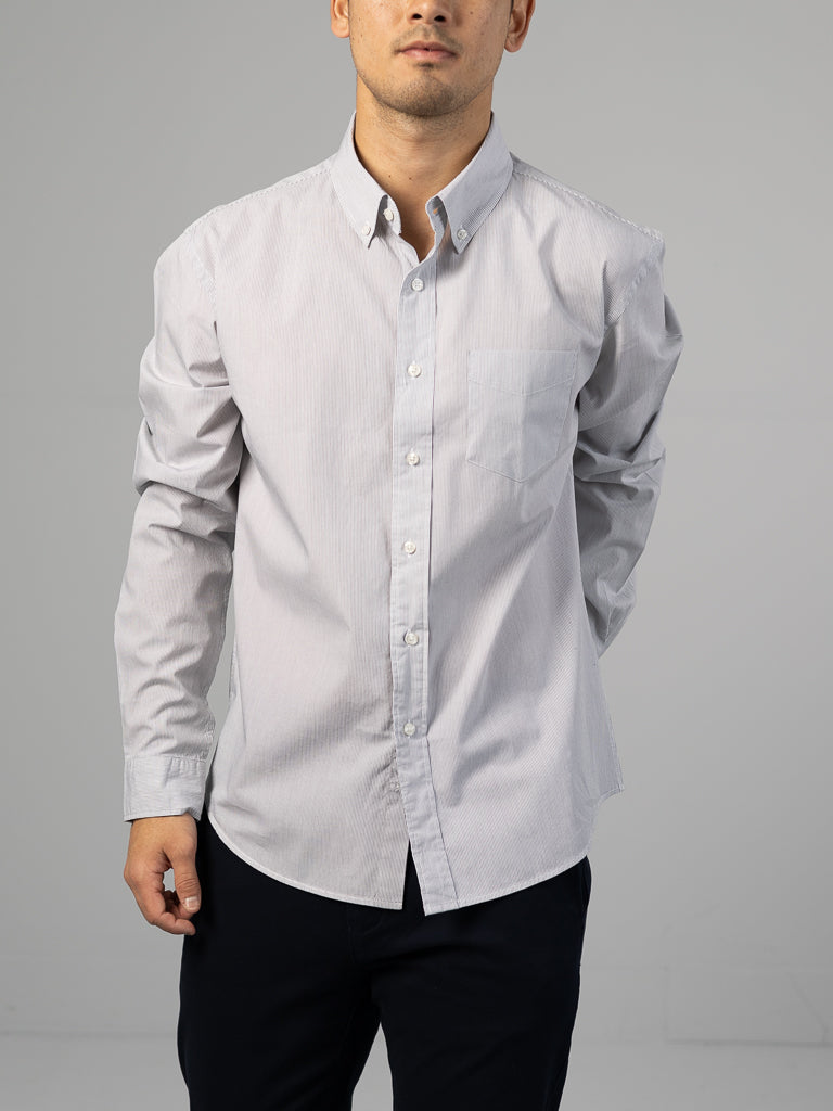 Someone wearing the ID Menswear Wade Stripe Button Down Shirt, a light gray, 100% cotton, regular-fit shirt with a left-side pocket, and dark pants stands against a plain gray background. Their face is not visible.