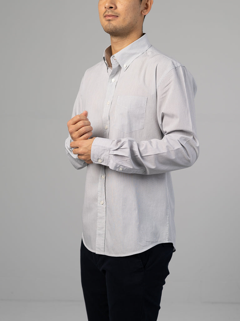 A person stands against a plain backdrop, dressed in an ID Menswear Wade Stripe Button Down Shirt and dark pants. They have short hair and their hands are clasped at their chest.