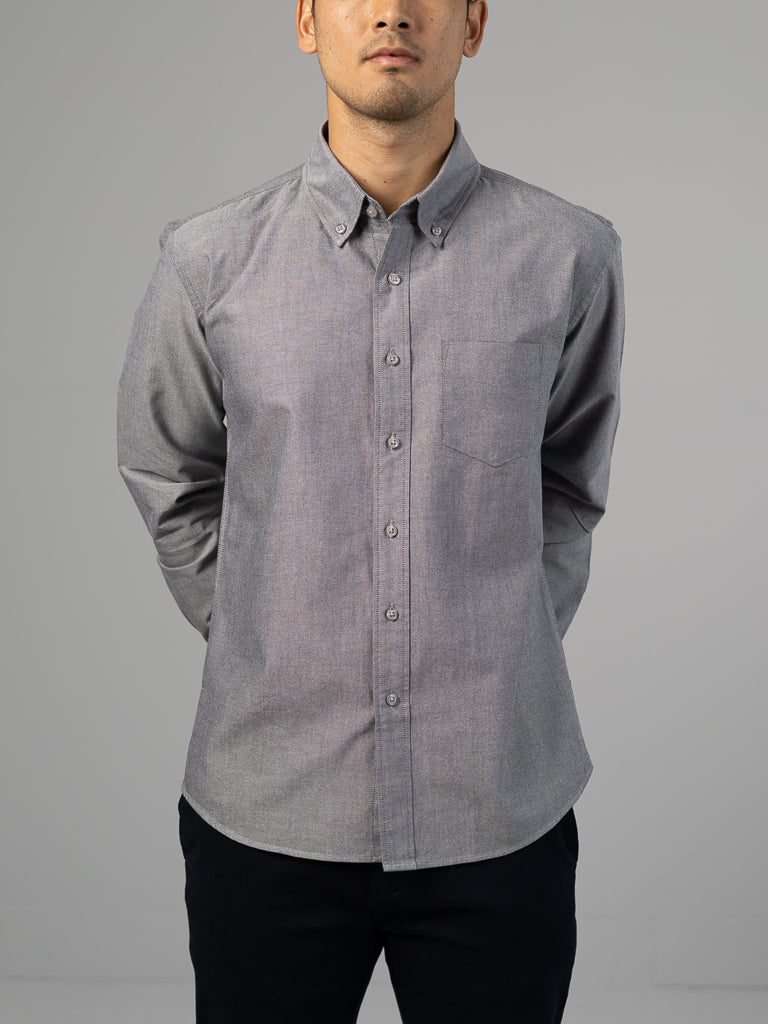 A person wears ID Menswears Oxford Long Sleeve Button Down Shirt in light gray, featuring a regular fit, pointed collar, and chest pocket. Made of 100% cotton, it pairs elegantly with dark pants as their hands rest behind their back against a neutral background.