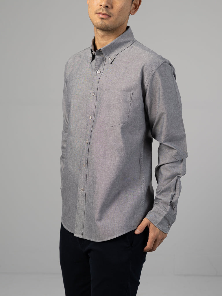 A person in a gray ID Menswear Oxford Long Sleeve Button Down Shirt and black pants stands against a gray background, the 100% cotton shirt offers a regular fit. With one hand in their pocket, they look slightly to the side.