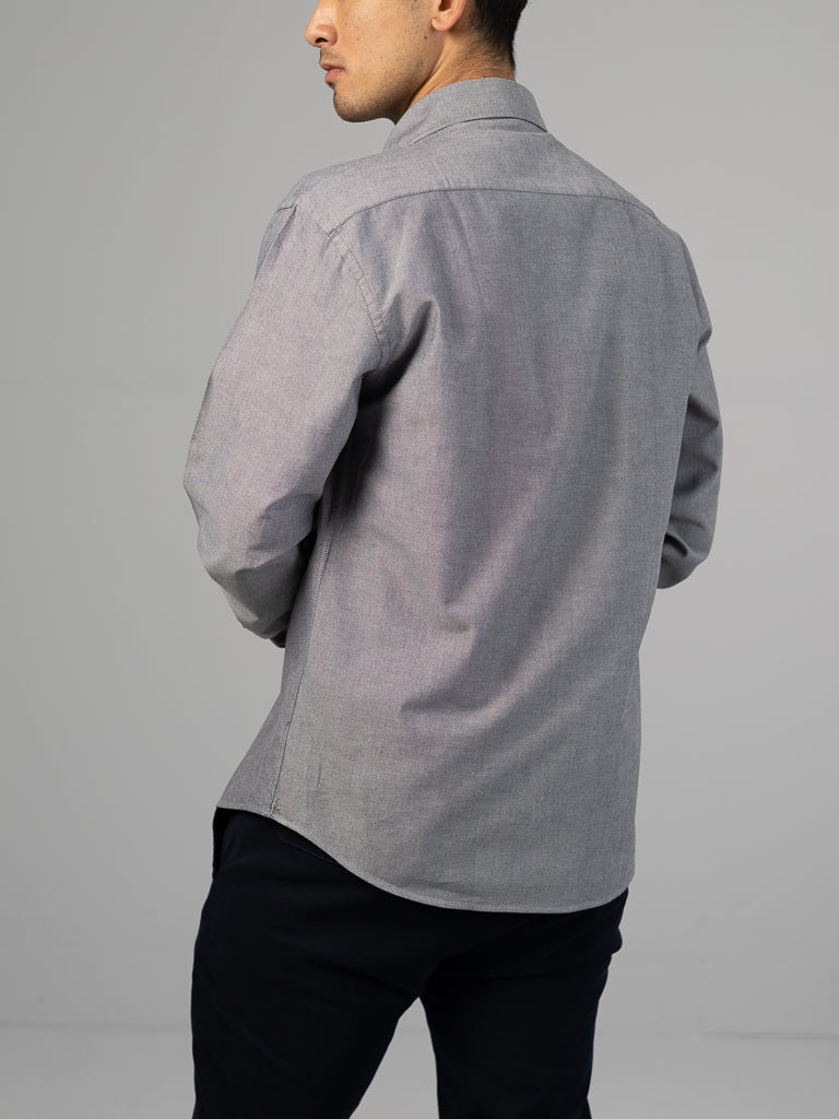 A person is standing slightly away from the camera, wearing an ID Menswear Oxford Long Sleeve Button Down Shirt in a regular-fit gray style and black pants, showcasing mostly their back and left side against a plain background. The 100% cotton fabric fits seamlessly into the scene.