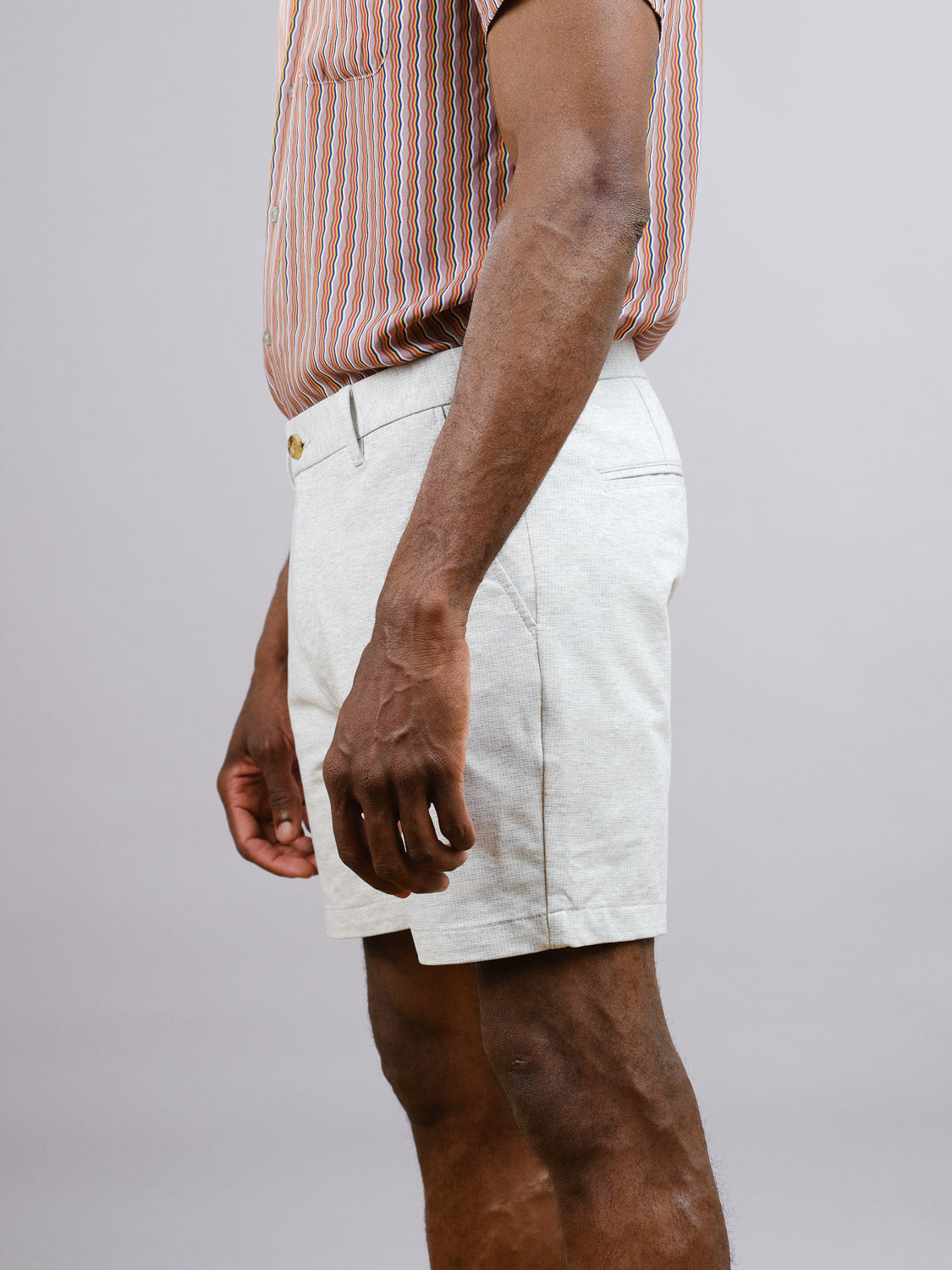 A person stands sideways, showing only their torso and upper legs, wearing a striped shirt tucked into light-colored ID Menswear Eckford 5 Inseam Stretch Shorts. The relaxed fit and comfort are visible against the plain background.