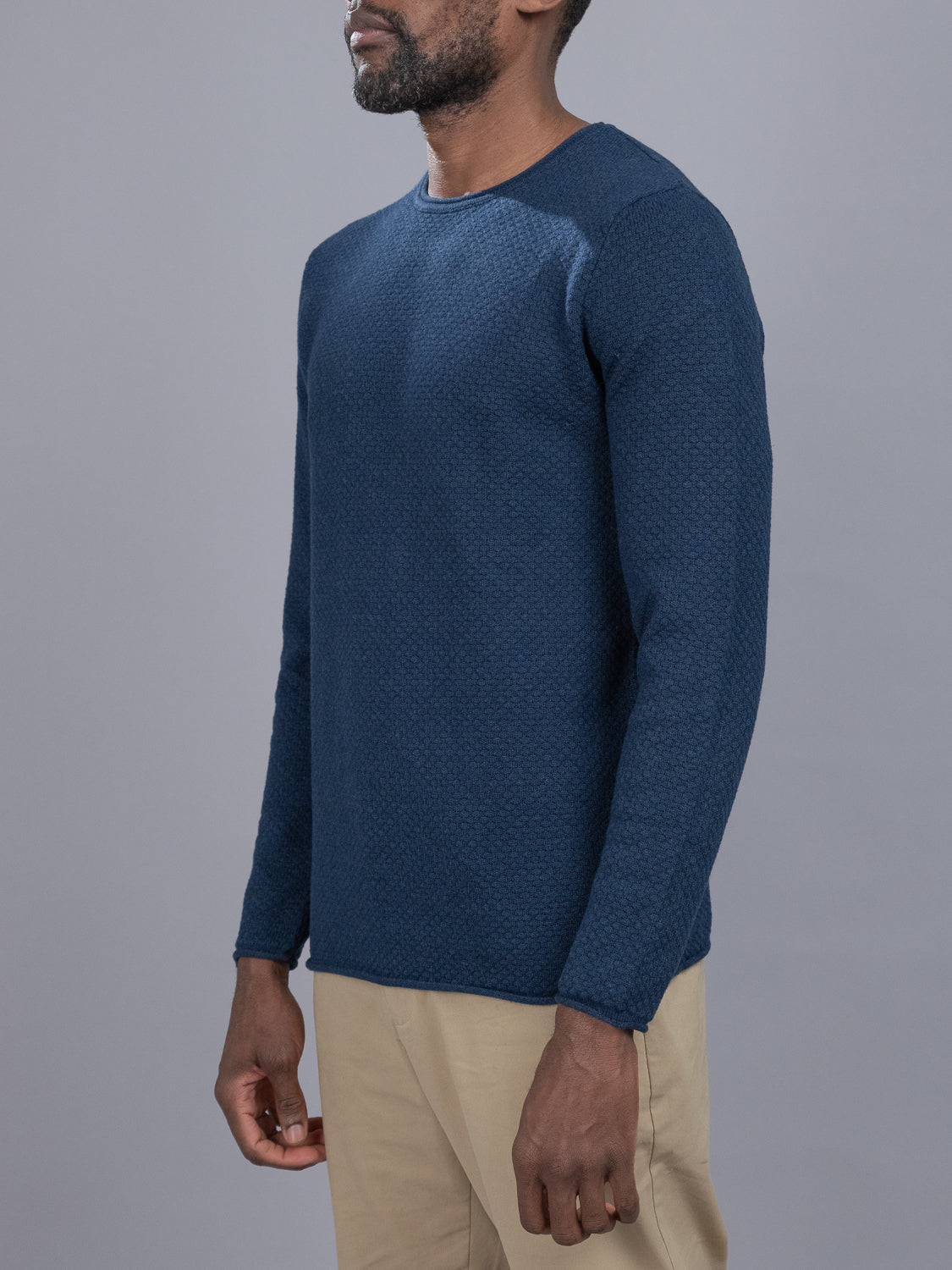 A man in an ID Menswear navy blue Westside Rolled Crew Neck Sweater and beige pants stands against a plain gray background, positioned at an angle with his hands by his sides.