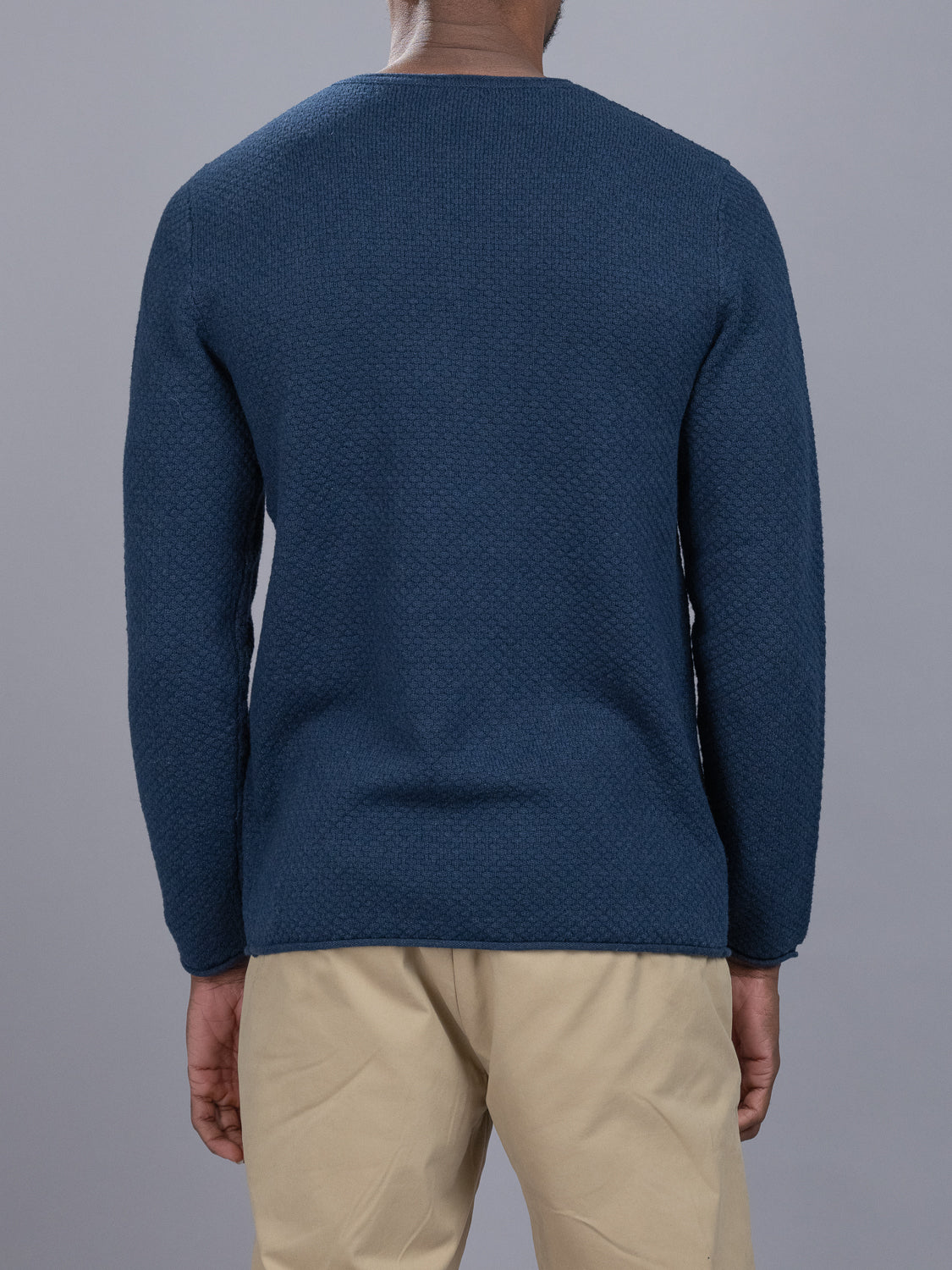 Rear view of a person wearing a textured dark blue ID Menswear Westside Rolled Crew Neck Sweater and beige pants, standing against a plain gray background.