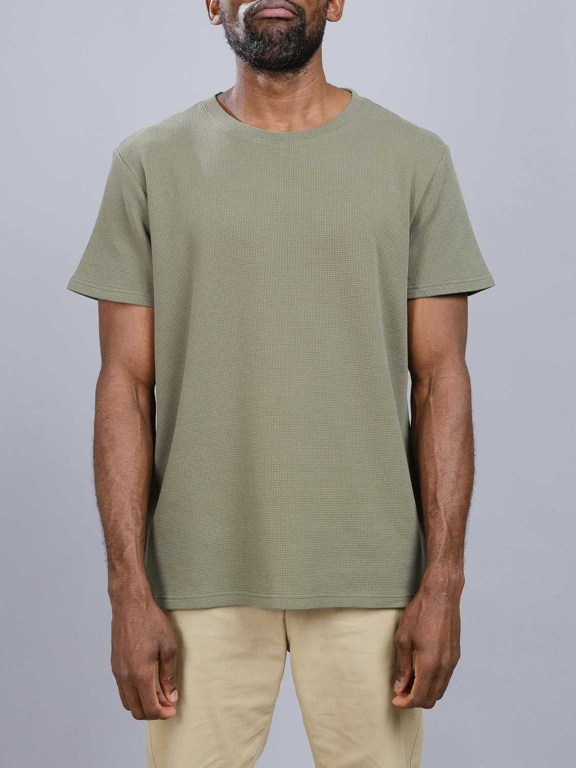 A person wears an olive-green Urban Waffle Knit Tee by ID Menswear and beige pants against a gray backdrop. Focused on the clothing, the classic tee offers comfort with its cotton-spandex blend. The persons head is partially out of frame.