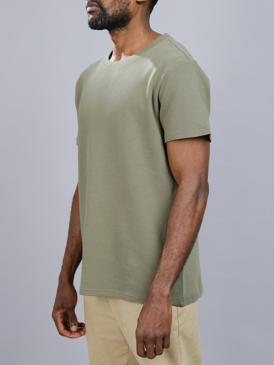 A person in an Urban Waffle Knit Tee by ID Menswear, a ribbed short-sleeved T-shirt in olive green, is paired with beige pants against a plain gray background. The cotton-spandex blend Tee is seen from the side, highlighting casual wear from shoulders to mid-thigh.